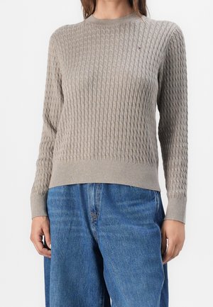 Femme portant un pull en maille torsadée beige à manches longues et un jean bleu taille haute, debout devant un fond blanc.