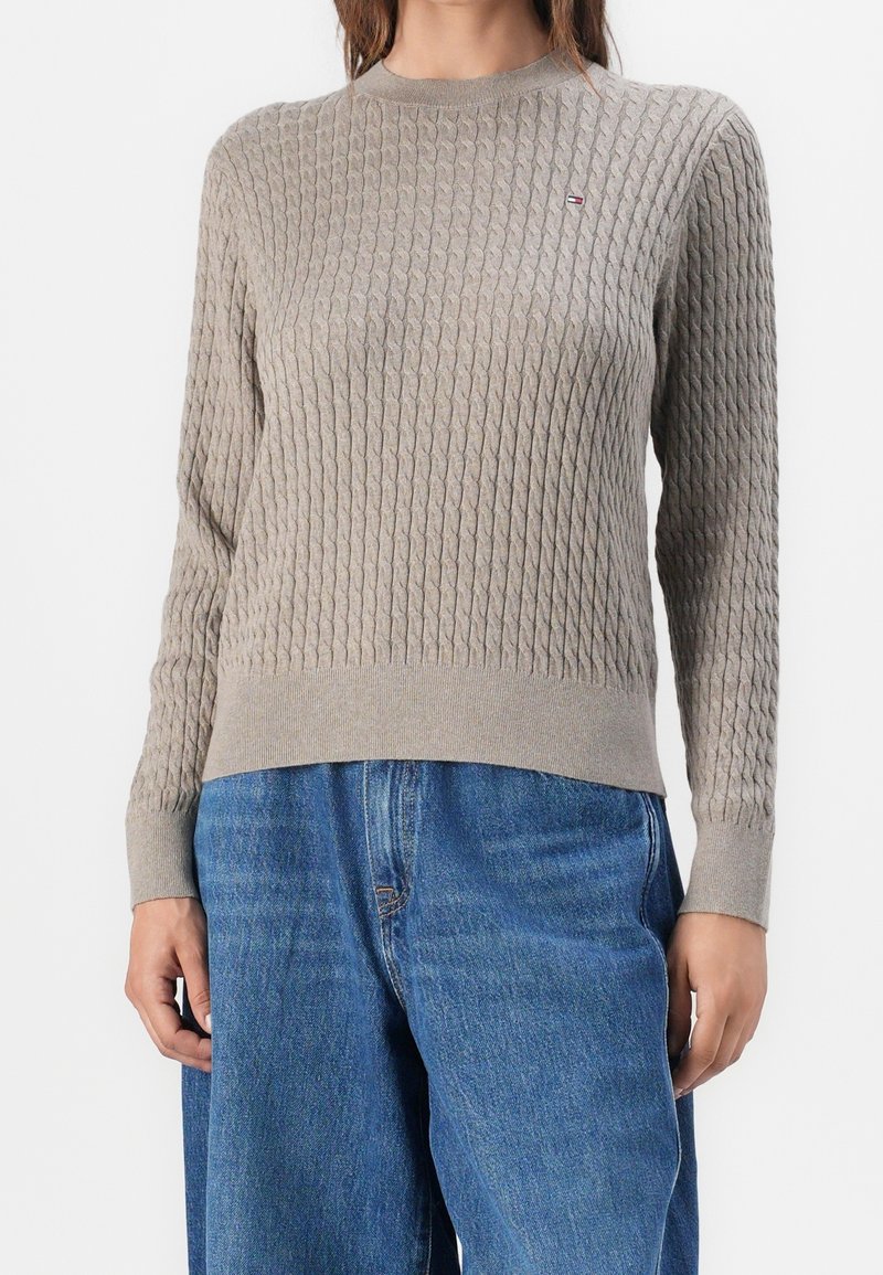 Femme portant un pull en maille torsadée beige à manches longues et un jean bleu taille haute, debout devant un fond blanc.