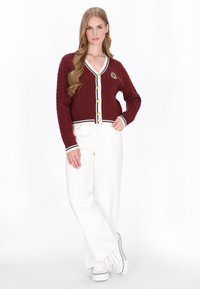 Burgundy cable-knit kardigan z głębokim dekoltem w kształcie litery V, złotymi guzikami, białym akcentem wykończenia i emblematem logo, w połączeniu z białymi spodniami typu wide-leg.