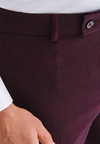 Zbliżenie osoby noszącej bordowe spodnie o fakturze, z guzikiem i szlufką na pasek, oraz białym rękawem koszuli widocznym po lewej stronie.