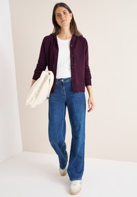 Cárdigan burdeos con botones, llevado sobre una camisa blanca, combinado con jeans de pierna ancha azules y zapatillas beige, sosteniendo un bolso crema.