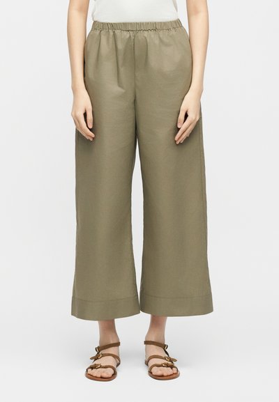 Γυναίκα που φοράει ελιά φαρδιά παντελόνια cropped με ελαστική μέση και καφέ φλατ σανδάλια με λουριά, στέκεται μπροστά σε ένα απλό φόντο.