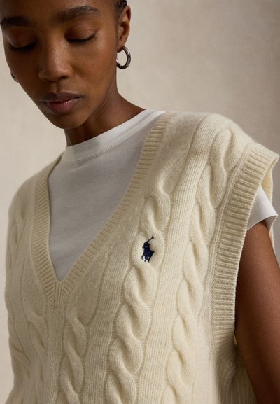 Zbliżenie osoby ubranej w biały, bezrękawnik w ściegiem warkoczowym na białej koszulce z małym granatowym logo na piersi.