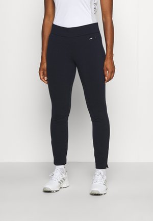 Mujer que lleva pantalones negros ajustados con bolsillo con cremallera, una camiseta blanca sin mangas y zapatillas deportivas blancas de pie sobre suelo gris.