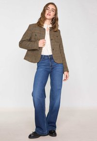 Vrouw in een bruine tweedjas, witte blouse met knopen, blauwe wijde jeans en zwarte schoenen, staand tegen een effen lichte achtergrond.