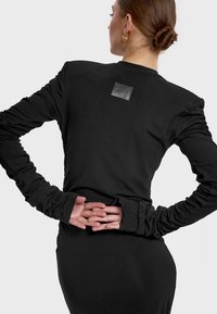 Czarna bluzka z długim rękawem, z marszczonymi rękawami i skórzaną naszywką z logo z tyłu; dopasowany krój z miękkiego, elastycznego materiału.