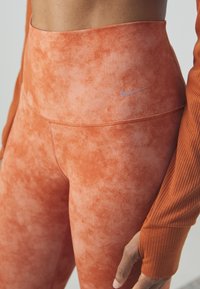 High-waisted leggings in a textured orange fabric, featuring a subtle gradient and a small, pink Nike logo on the waistband.