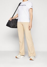 Person i vit Champion T-shirt och beige byxor, håller en svart Puma gymväska över axeln, står framför en enfärgad bakgrund.