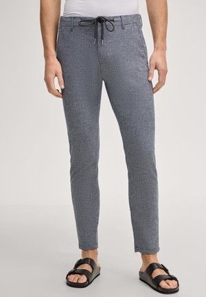 Uomo con pantaloni grigi slim-fit con coulisse e sandali neri con fibbia, in piedi davanti a uno sfondo chiaro.