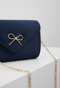 Sac à main en satin bleu marine avec un accent en nœud doré et une chaîne. Le design est structuré avec une fermeture à rabat et une texture lisse.