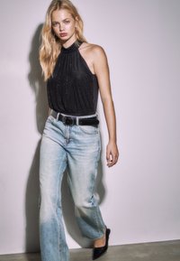 Zwarte geplooide haltertop met subtiele glans, gecombineerd met lichtblauwe wijd uitlopende jeans en zwarte pumps met een puntige neus. Effen achtergrond.