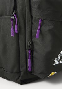 Mochila preta com tecido texturado, apresentando puxadores de zíper roxos e ferragens pretas. Inclui um logotipo branco em destaque e uma etiqueta de destaque colorida.