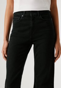 Fekete farmernadrág, amely egyenes szabású, öt zsebes kialakítással, kontrasztos varrással és gombos záródással rendelkezik. Az anyag texturált és tartós hatású.