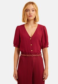 Bordeauxfarbene Bluse mit V-Ausschnitt, Puffärmeln und goldenen Knopfdetails. Kombiniert mit passender, plissierter Hose und einem goldenen Kettenbelt.