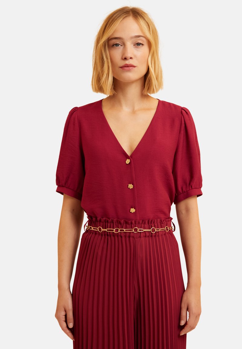 Bordeauxfarbene Bluse mit V-Ausschnitt, Puffärmeln und goldenen Knopfdetails. Kombiniert mit passender, plissierter Hose und einem goldenen Kettenbelt.
