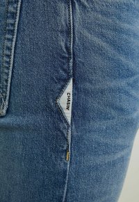 Gros plan sur une poche de jean en denim bleu avec une étiquette triangulaire blanche affichant le nom de la marque "CHASIN" en lettres noires.