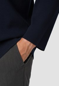 Marineblauwe pullover met geribbelde mouwen, gecombineerd met grijze broek. De pullover heeft een getextureerde brei, met de hand in de zak die de materialen en het ontwerp benadrukt.