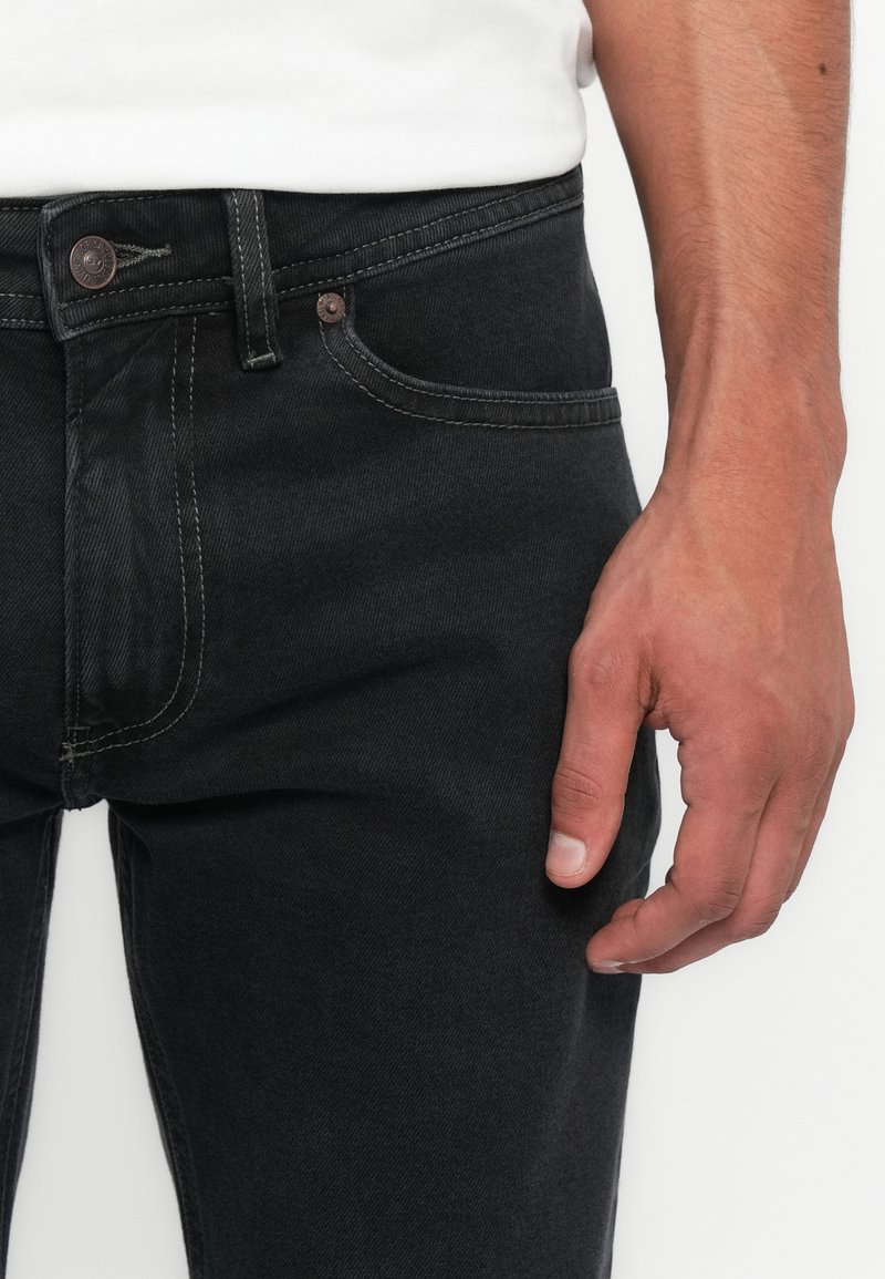 Nahaufnahme einer Person, die schwarze Jeans und ein weißes Hemd trägt, mit Fokus auf der Taille, dem Knopf, den Taschen und der rechten Hand.