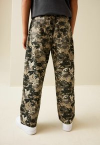 Pantaloni cargo cu model camuflaj în nuanțe de khaki și verde închis, având buzunare laterale și un croi lejer. Pantalonii sunt asortați cu adidași albi.