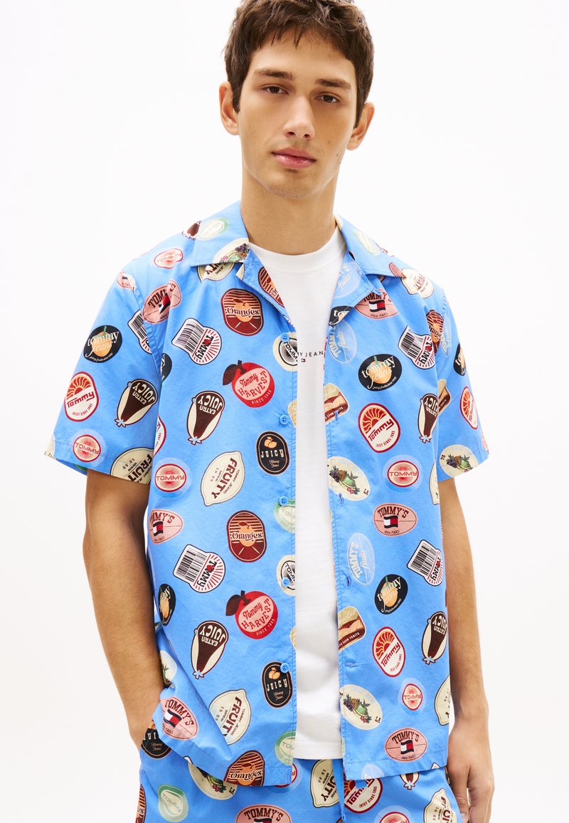 Ung mand iført lyseblå kortærmet skjorte og shorts med farverige vintage madlabel-mønstre, stående mod en hvid baggrund.