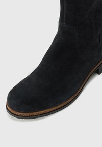 Bottine en suede noir avec un bout rond, des coutures détaillées et une semelle brune contrastante. Surface texturée et finition lisse.