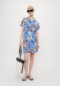 Vestido de mangas curtas com um padrão floral em azul, preto e creme, cintura ajustada e colarinho. Modelo segura uma bolsa tote preta e creme.