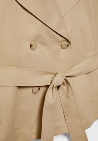 Trench-coat beige à double boutonnage avec quatre boutons et une ceinture en tissu nouée à la taille.