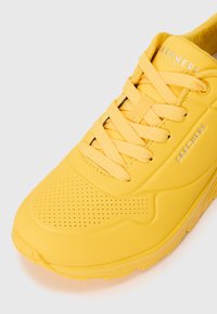 Gelbe Skechers Sneaker mit einem perforierten Design, flachen Schnürsenkeln und einer strukturierten Gummisohle. Die Markenbezeichnung ist in Silber an der Seite sichtbar.
