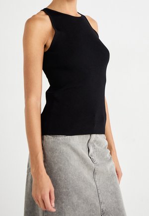 Femme portant un haut noir côtelé sans manches et une jupe en jean gris clair sur un fond blanc uni.