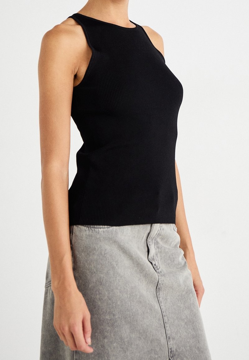 Femme portant un haut noir côtelé sans manches et une jupe en jean gris clair sur un fond blanc uni.