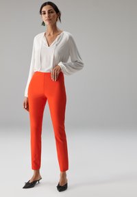 Blouse blanche à manches amples, col en V profond ; pantalon orange ajusté ; mules noires avec détail nœud ; tissu texturé, coupe cintrée.