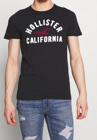 Camiseta de algodón negra que presenta "Hollister California" en letras blancas y rojas, con cuello redondo, mangas cortas y un diseño ajustado.