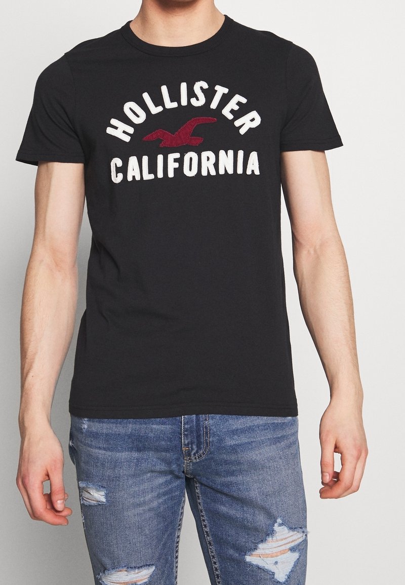 Camiseta de algodón negra que presenta "Hollister California" en letras blancas y rojas, con cuello redondo, mangas cortas y un diseño ajustado.