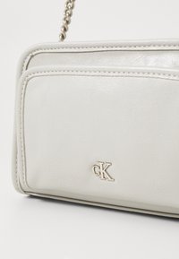 Bílá kabelka přes rameno z umělé k�ůže se zlatými kovovými prvky, která má jemně embosované logo a prošívané akcenty. Součástí je odnímatelný řetízkový popruh.