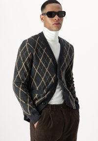 Mørkegrønn og grå argyle cardigan med beige detaljer, ribbestrikket krage og mansjetter, knappelukking, kombinert med hvit høyhalset genser og brune kordfløyelsbukser.
