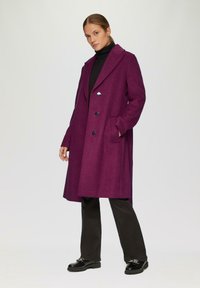 Fuchsia wollen jas met een op maat gemaakt ontwerp, grote kraag, dubbele knopen aan de voorkant en twee zakken, gecombineerd met zwarte broeken en schoenen.