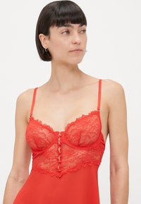 Bralette en dentelle de couleur rouge vif avec bretelles ajustables, motif floral et boutons décoratifs à l'avant central, fabriqué en tissu transparent et lisse.