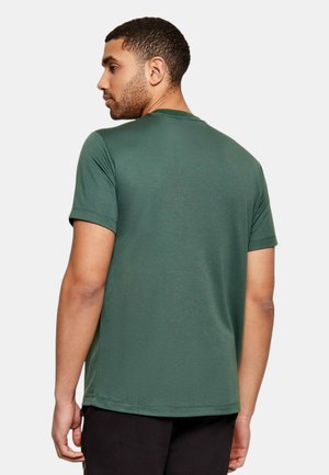 Uomo con capelli corti e ricci visto di spalle, che indossa una t-shirt verde scuro a maniche corte e pantaloni neri, su sfondo grigio.