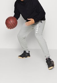 Šedé tepláky s nápisem "PARIS" vytištěným bílou barvou, v kombinaci s černými teniskami a tmavou mikinou na zip. Basketbalová míč je v popředí.