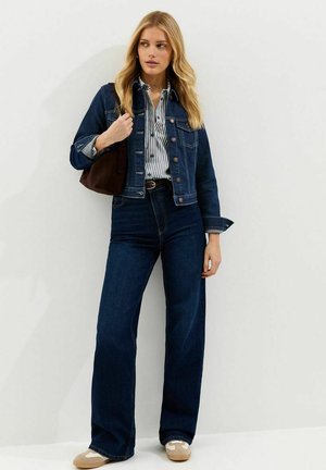 Kobieta w ciemnej dżinsowej kurtce i dżinsach, w paskowaną koszulę, beżowe sneakersy, z brązową torbą na ramię, stoi na tle białej ściany.