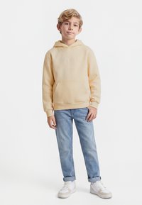 Blondiem zēns ar bēšas krāsas kapuci, gaiši zilām sarullētām džinsa biksēm un baltām sporta kurpēm, stāv ar atslābinātām rokām.