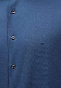 Μπλε πουκάμισο με κουμπιά και τρία σκοτεινά κουμπιά, καθώς και μικρό κεντημένο λογότυπο "MK" στη δεξιά πλευρά του στήθους.