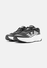 Fekete atlétikai cipők texturált felsőrésszel, fehér, geometrikus középtalppal és fekete gumitalppal; jellegzetes logóval és csíkdíszítéssel.