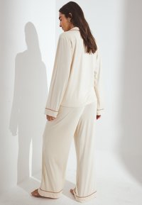 Beige pidžamu komplekts mīkstā audumā, kuram ir garas piedurknes un platai bikses, ar kontrastējošiem apdari uz piedurknēm un biksēm.