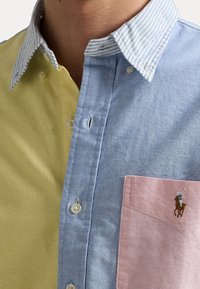 Wielokolorowa koszula z żółtymi, niebieskimi i różowymi sekcjami; w paski kołnierzyk. Posiada guziki i małe haftowane logo na kieszeni na piersi.