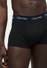 Ανδρικός κορμός με μαύρα μποξεράκια Calvin Klein, εμφανίζοντας τη ζώνη με το λογότυπο Calvin Klein και έναν μυώδη βραχίονα στο πλάι.