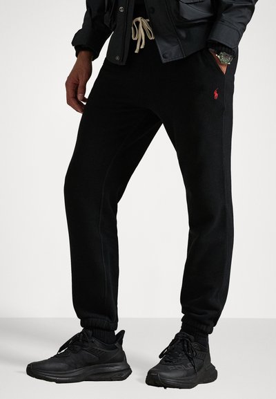 Zwarte katoenen sweatpants met elastische boorden, voorzien van een rood logo, een koordsluiting in de taille en zijzakken. Gemaakt in combinatie met zwarte sport schoenen.