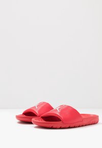 Punaiset liukuuvaiset sandaalit, joissa on teksturoitu päällinen ja valkoinen logo. Pohja on tasainen ja siinä on aaltoilu, joka tarjoaa pidon.