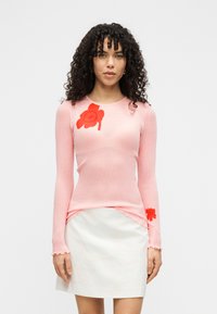 Ριμπ μπλουζάκι με μακριά μανίκια σε ανοιχτό ροζ χρώμα με κόκκινη κεντητική με σχέδιο λουλουδιών στο στήθος και το μανίκι· συνδυάζεται με λευκή κοντή φούστα.