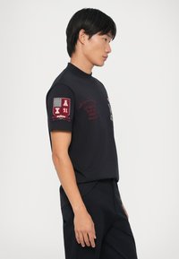 T-shirt nera a maniche corte con collo alto, caratterizzata da ricami rossi sulle maniche e sul davanti. Realizzata in un tessuto liscio, ha un design aderente.